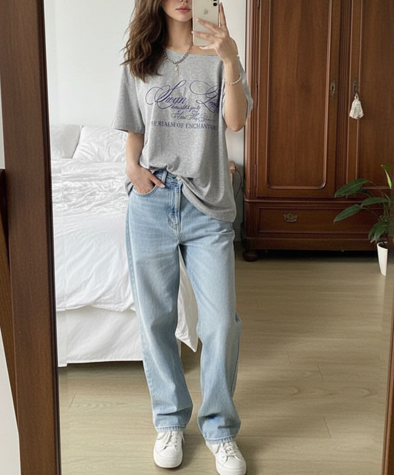 Person taking a mirror selfie wearing a gray t-shirt and light blue jeans in a bedroom.